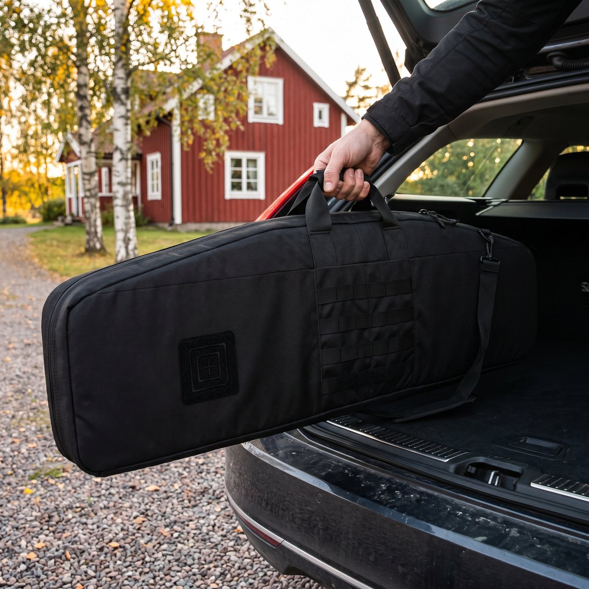 Svart 5.11 Single Rifle Case lyfts i topphandtaget in i bagageutrymmet vid ett rött svenskt trähus