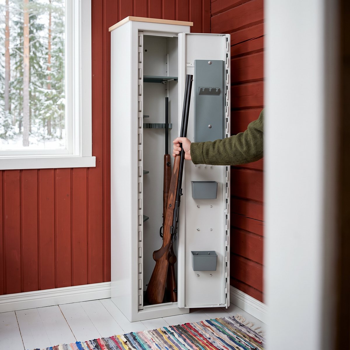 Smalt, högt ljust vapenskåp Nor-Lyx HL5 med träskiva på toppen i en hall med rödmålade väggar, där en hand ställer in ett jaktgevär