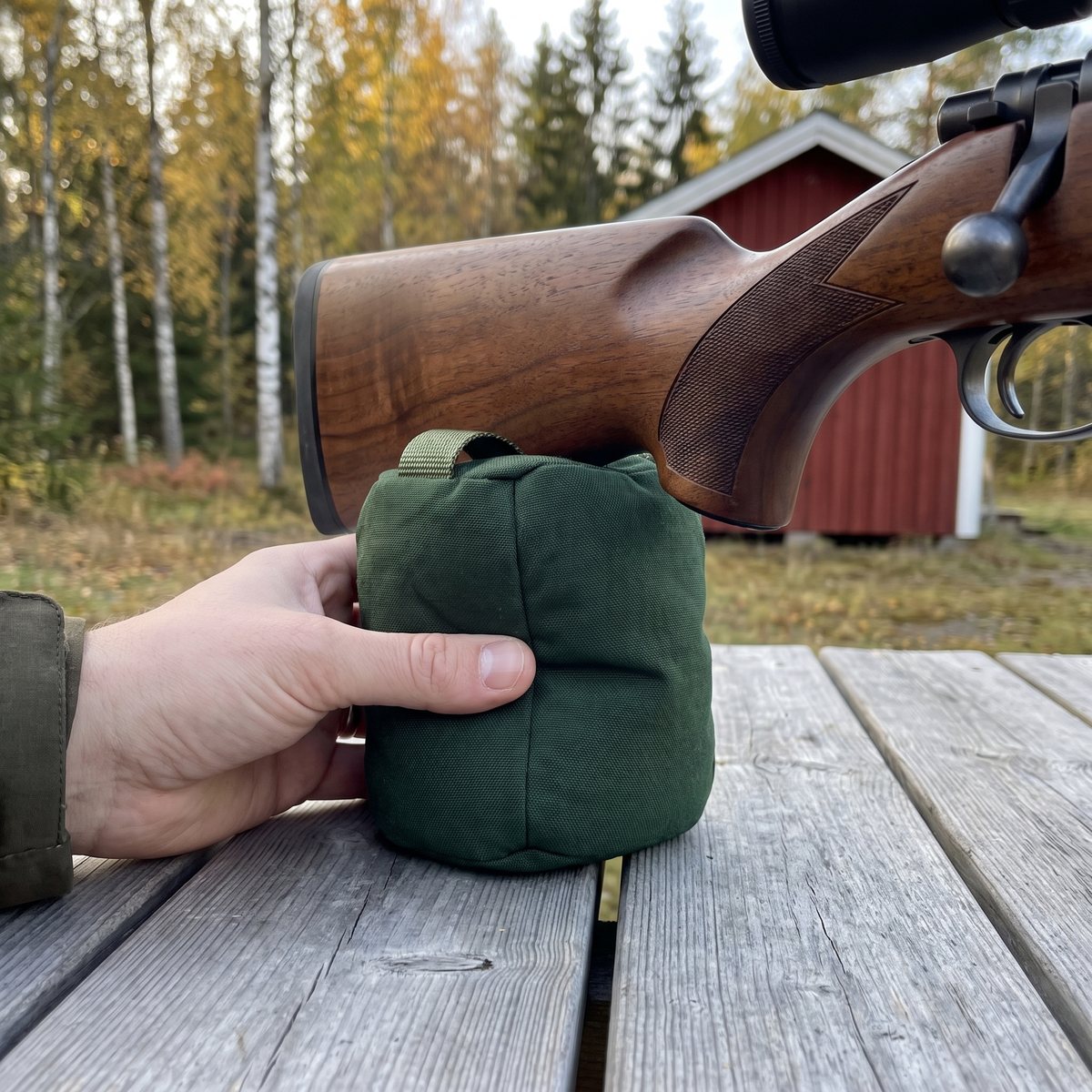 Mörkgrön baksäck under kolven på ett jaktgevär med valnötsstock på ett skjutbord, med rött uthus och björkar i bakgrunden