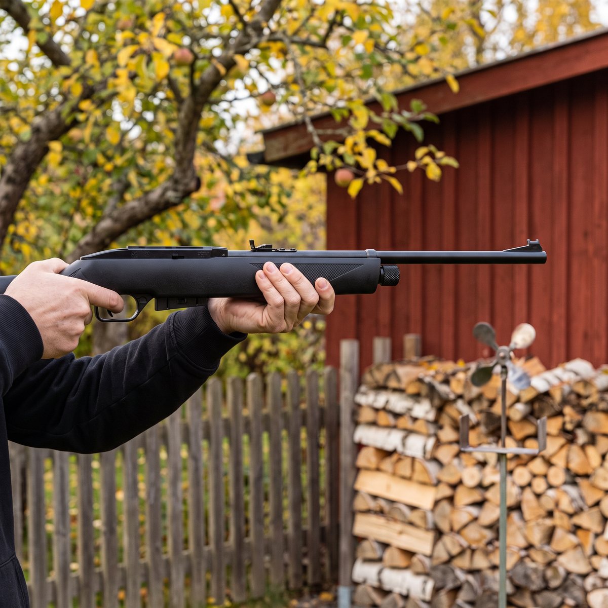 Crosman 1077 i stående skytte mot en snurrmåltavla i en svensk trädgård med vedtrave och rött förråd