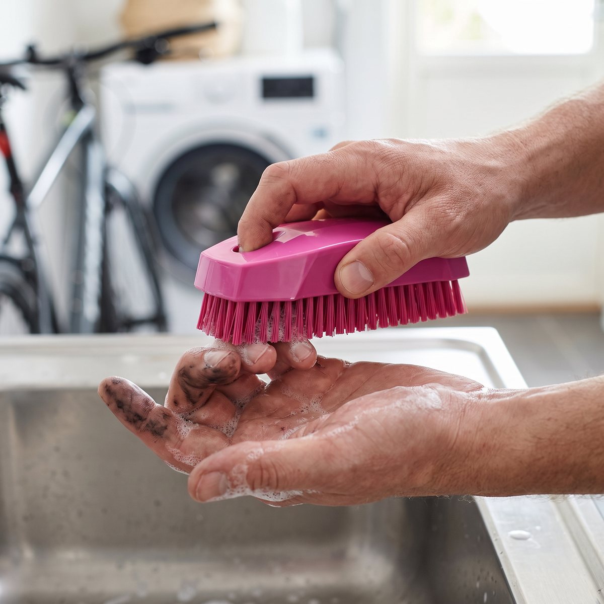 Hand skrubbar cykelolja från fingrarna med en rosa nagelborste över en diskho i en svensk tvättstuga