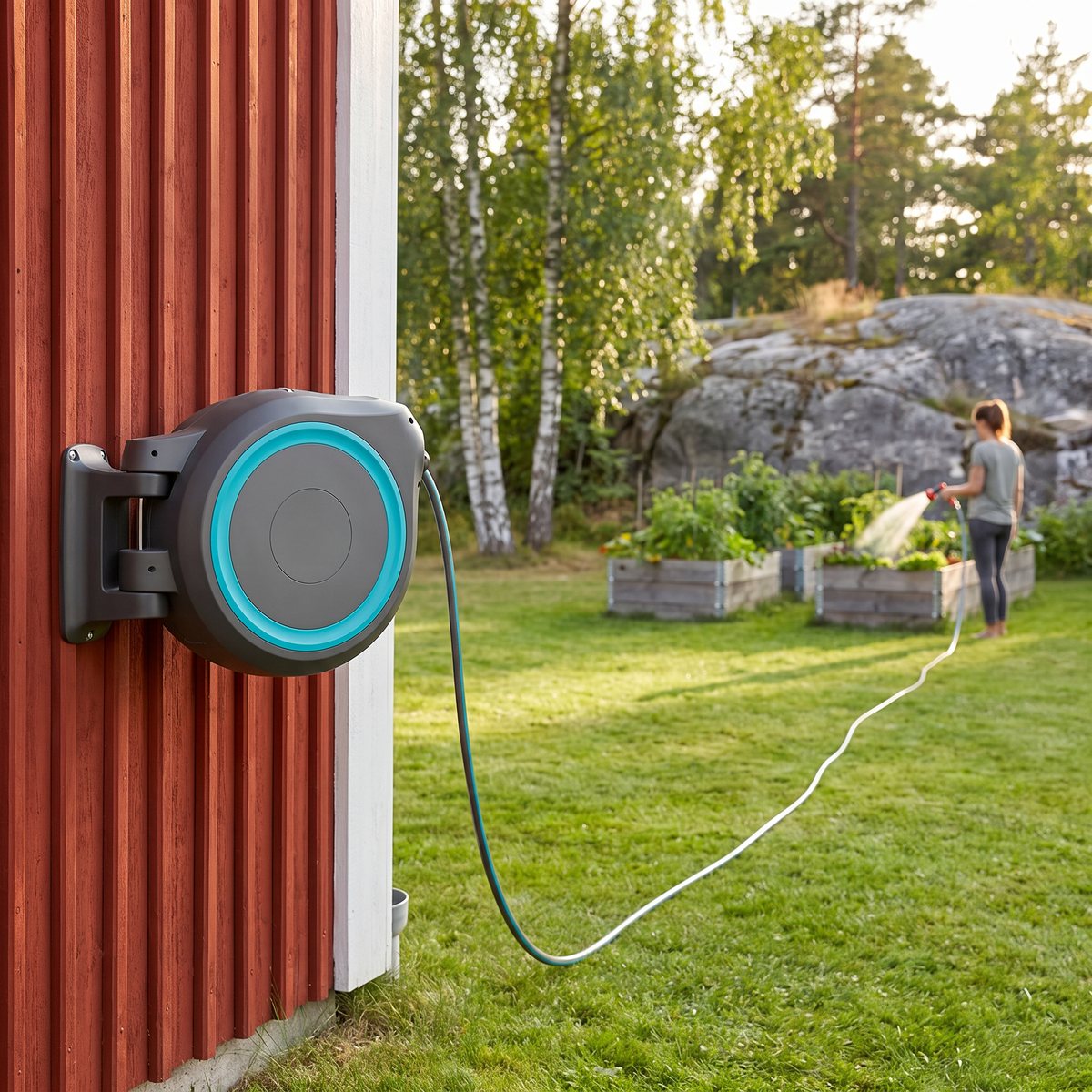 Gardena RollUp XL monterad på en falurödmålad husvägg, med slangen utdragen över gräsmattan till odlingslådor där någon vattnar