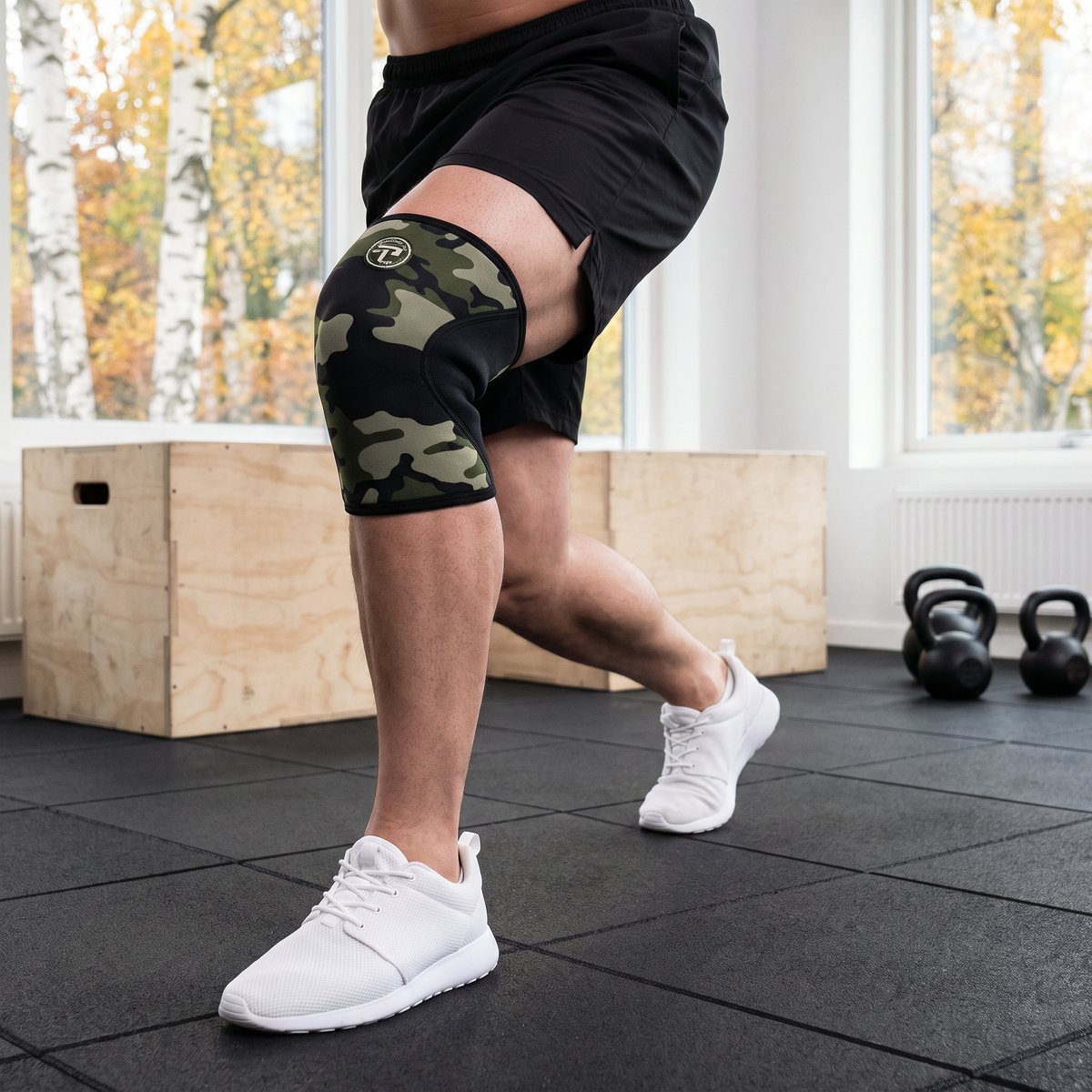 Kamouflagemönstrad neoprenhylsa på knät hos en person som gör utfallssteg i ett ljust svenskt träningsrum med plyoboxar och kettlebells