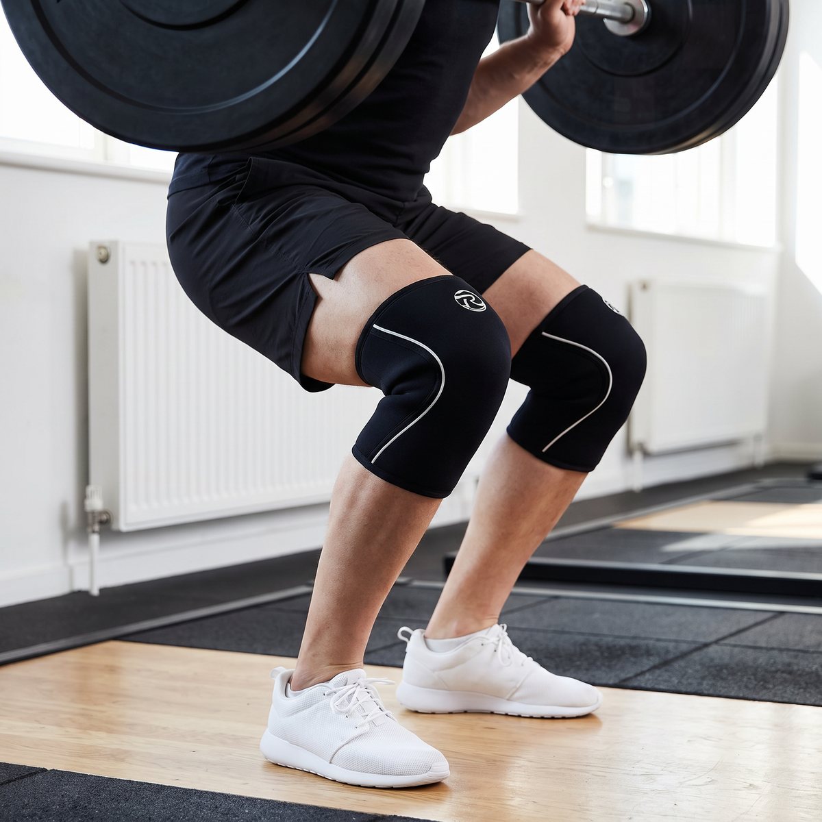 Svarta neoprenhylsor med vita sömmar på båda knäna i botten av en tung knäböj med skivstång i ett svenskt gym