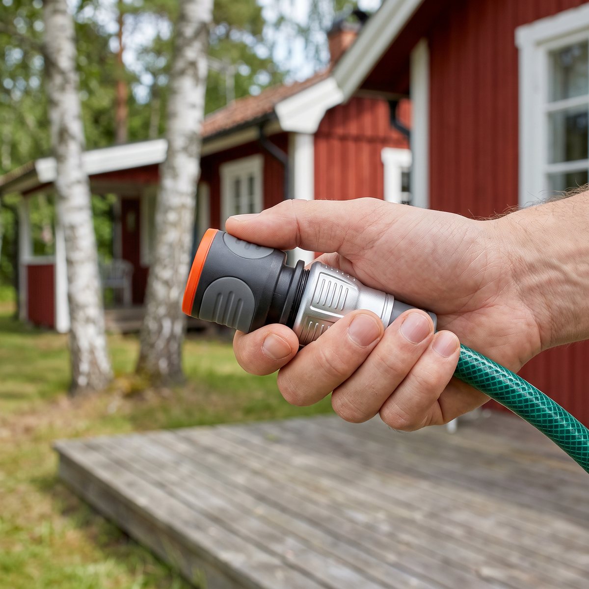 Gardena Premium slangkoppling kopplad på en trädgårdsslang, hållen i handen vid en röd stuga med björkar