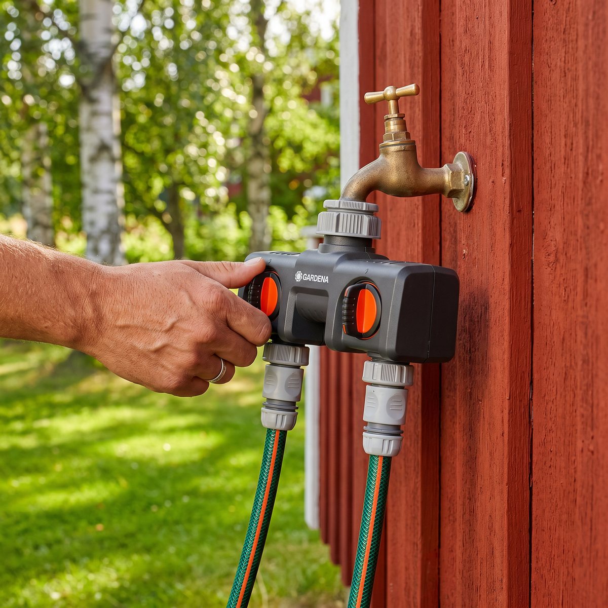 Gardena vattenfördelare med två uttag monterad på en utomhuskran på en röd trähusvägg, med en hand på ett av de orange reglagen