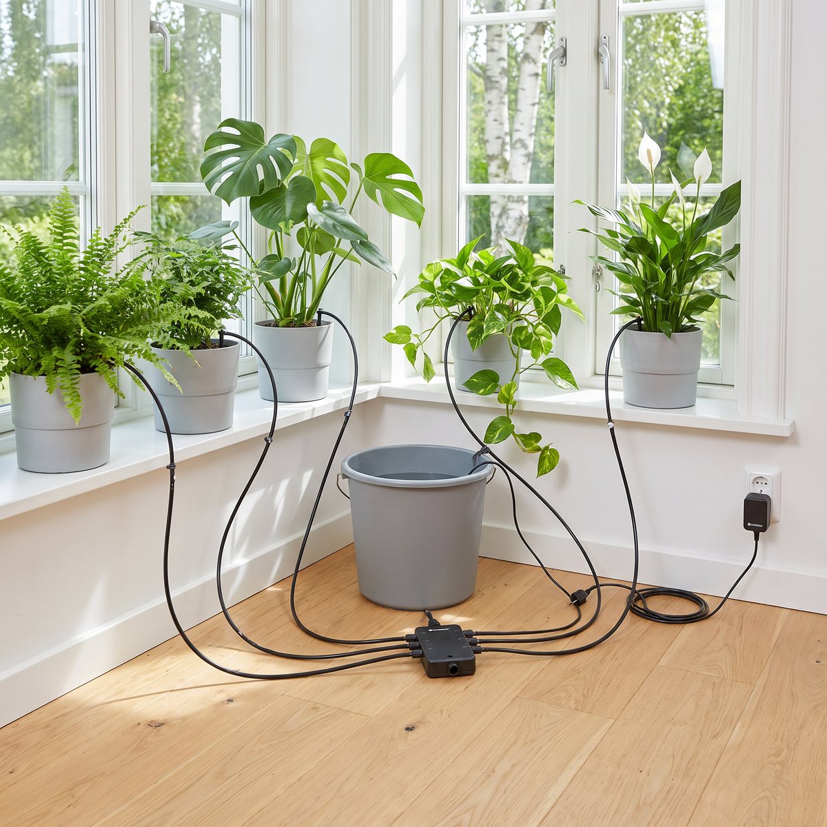 Gardena City Gardening med droppslangar till krukväxter i ett ljust fönster, vattenhink på golvet och nätadapter i vägguttaget
