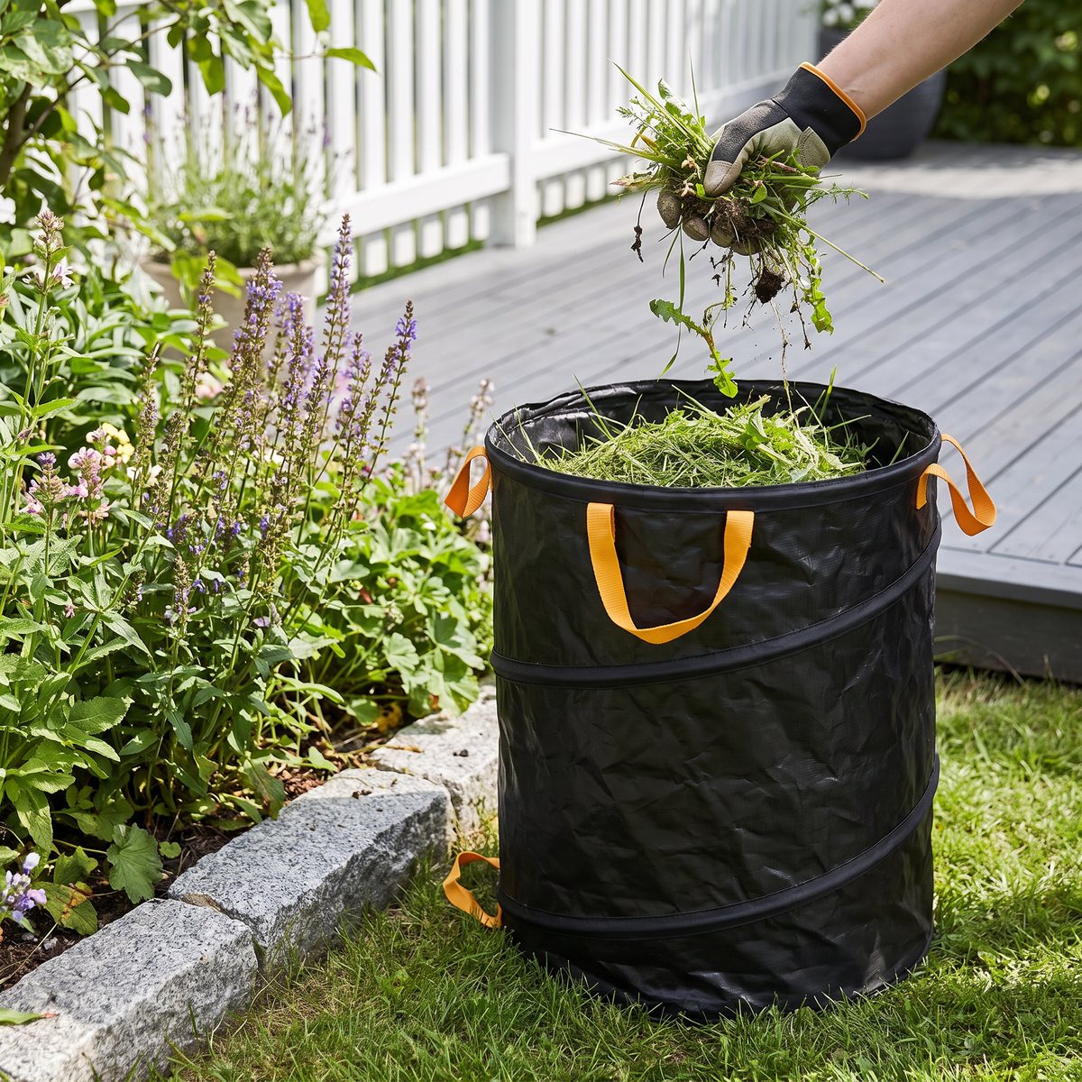 Fiskars Pop Up Solid, en svart rund trädgårdssäck med orange handtag, bredvid en rabatt med gräsklipp och ogräs