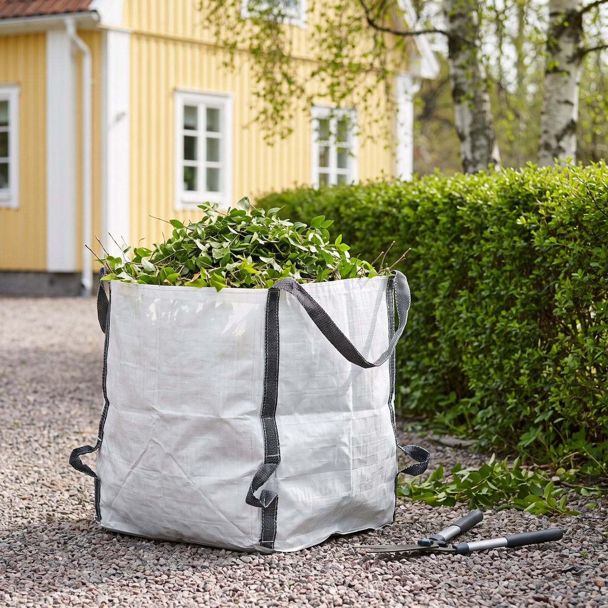 Vit vävd trädgårdssäck Nelson Garden WasteBag fylld med häckklipp på en grusgång bredvid en klippt häck
