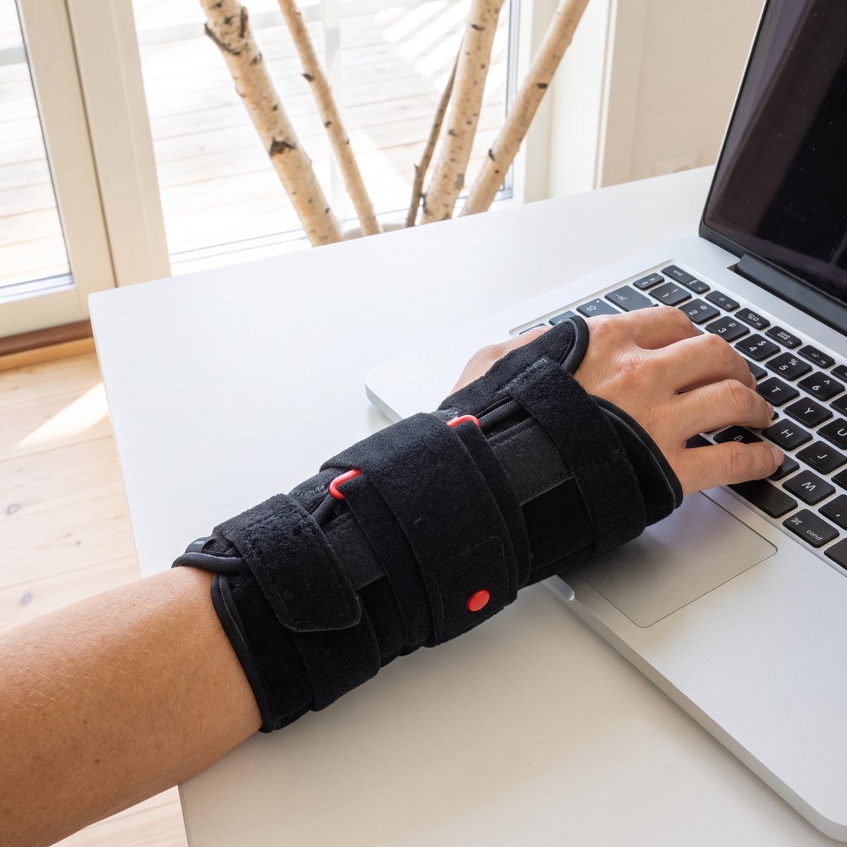 Svart handledsskena med kardborrband och röda detaljer på en hand som skriver på laptop i svenskt hem