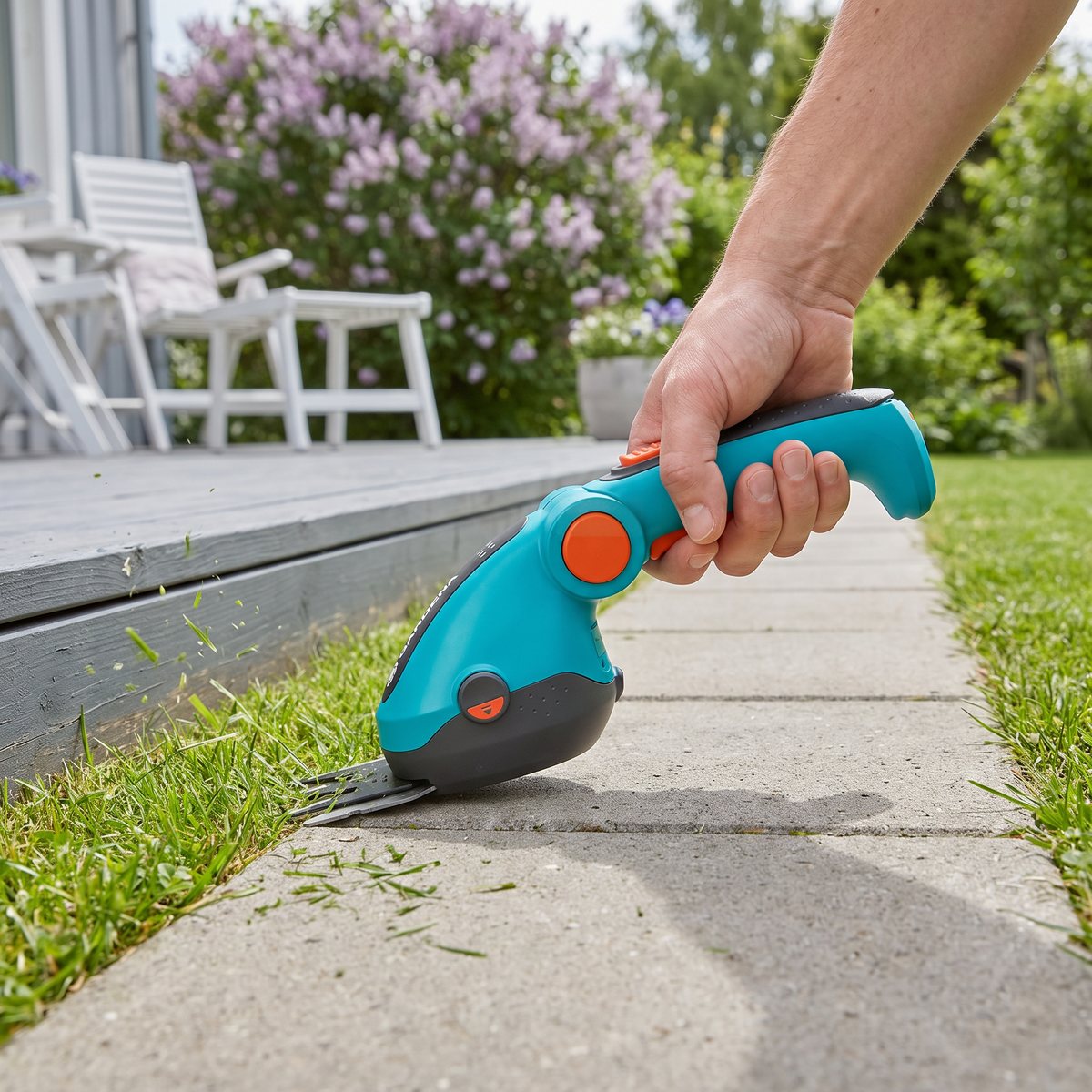 Gardena ComfortCut Li batteridriven grässax klipper gräskanten längs en plattsatt gång vid ett grått trädäck med syren i bakgrunden