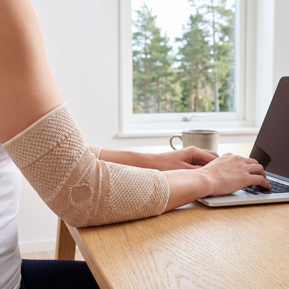 Beige stickat armbågsbandage på en underarm vid ett skrivbord i ett svenskt hemmakontor