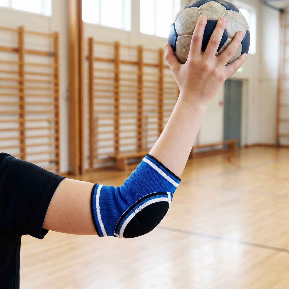 Blått armbågsskydd för junior på en arm som håller en handboll i en svensk skolgympasal