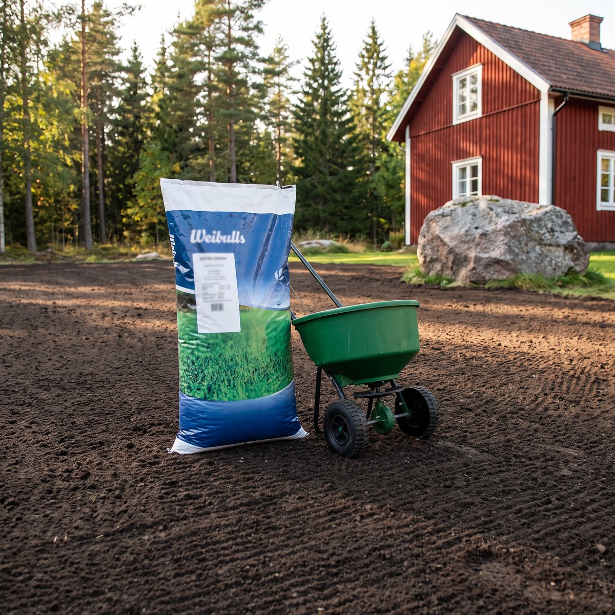 Säck med Weibulls Extra Green bredvid en fröspridare på en stor krattad jordyta vid ett rött hus och granskog
