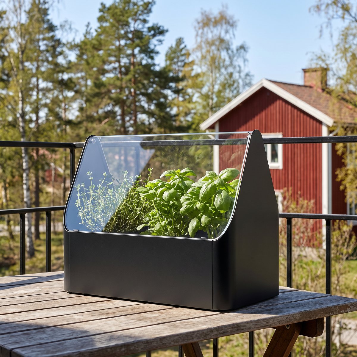 Eva Solo Simply Grow med basilika och timjan bakom glaset, på ett balkongbord med tallar och ett rött trähus i bakgrunden