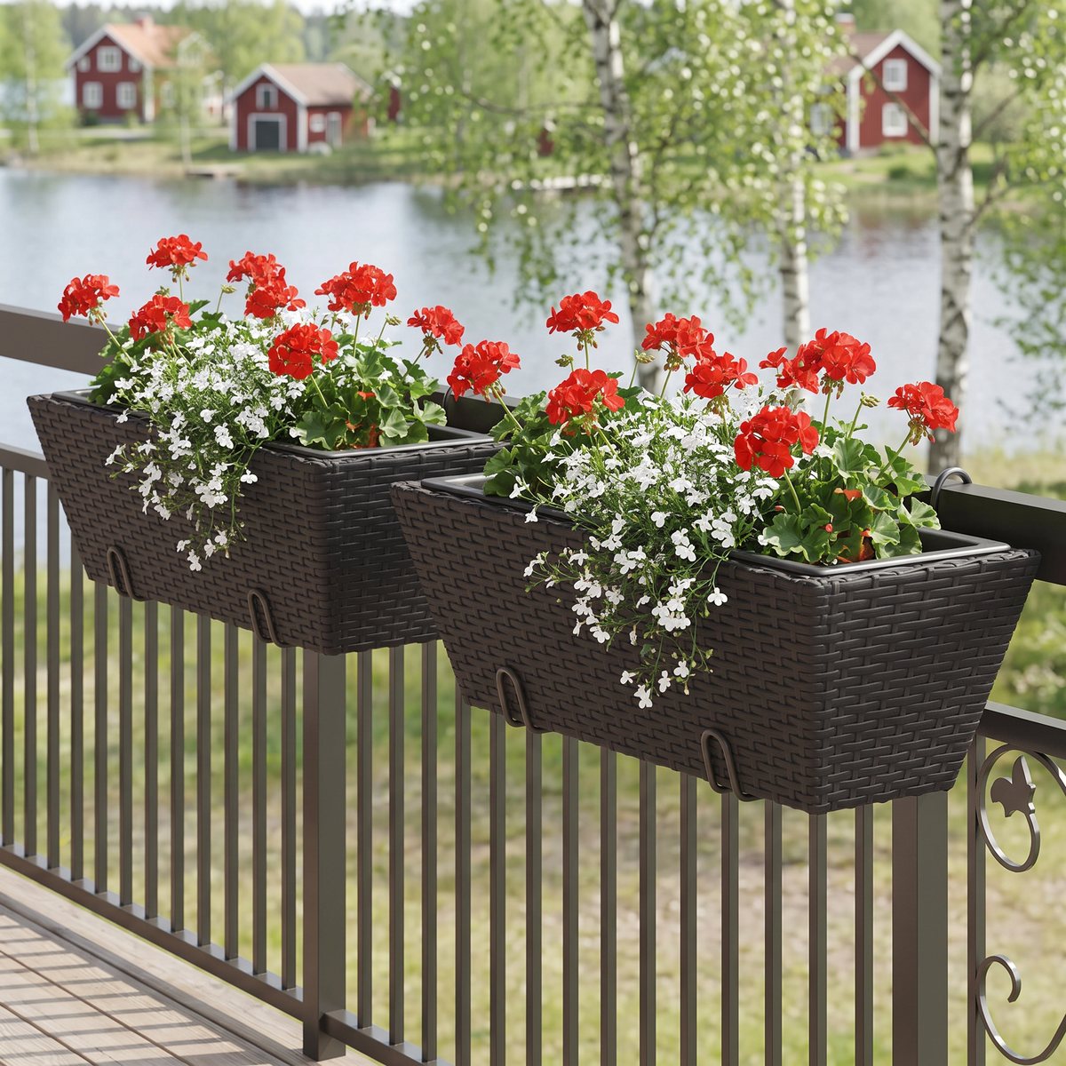 Två svarta blomlådor i konstrotting hänger med krokar på ett balkongräcke, planterade med röda pelargoner och vit lobelia, med en svensk sjö och röda trähus i bakgrunden