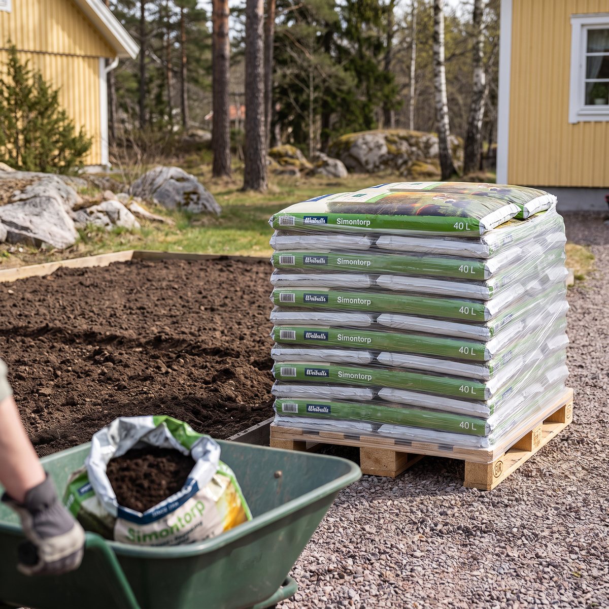 Plastad helpall med säckar Weibulls Simontorp Kogödsel på en grusuppfart bredvid ett nygrävt grönsaksland, med en öppnad säck i en skottkärra, gult trähus, tallar och granithällar