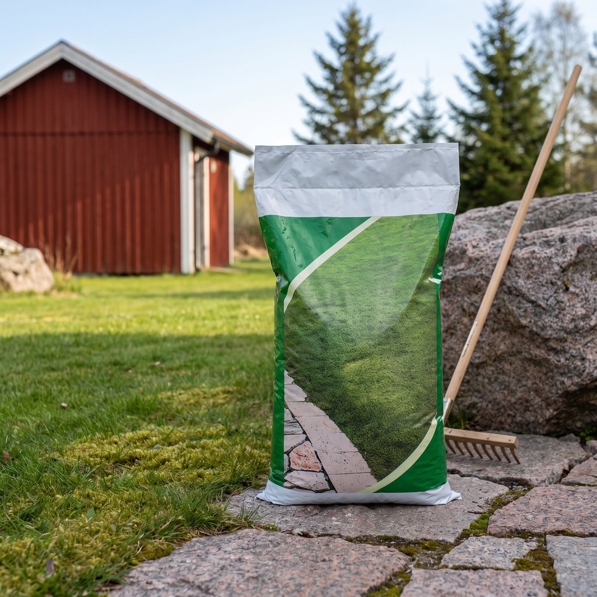 Säck med Greenkeeping Gräsgödsel Mossa & Ogräs på en stenplatta intill en gräsmatta med kratta och rött skjul