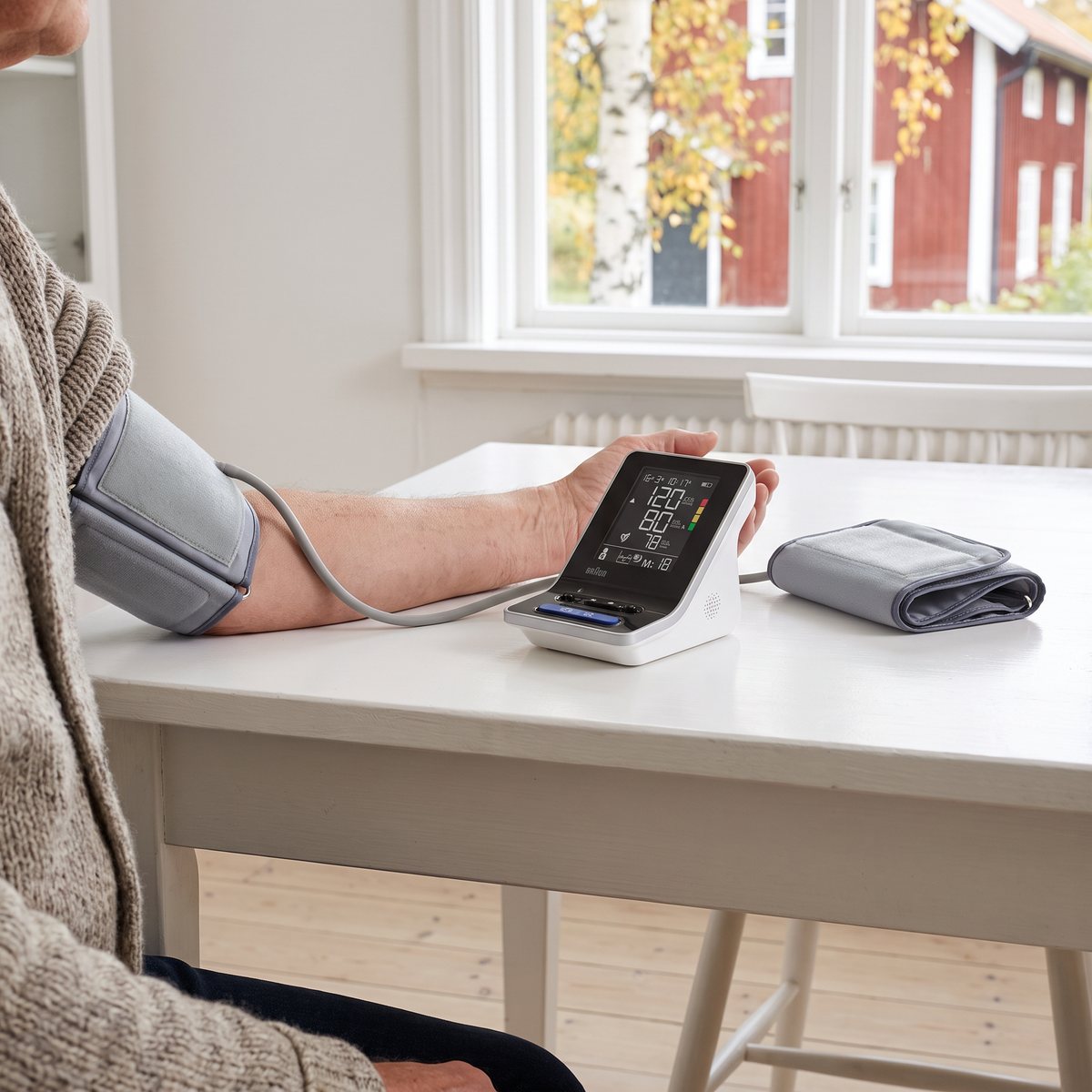 Braun ExactFit 3 på ett vitt matbord med tygmanschetten på överarmen och en extra manschett bredvid, rött trähus utanför fönstret
