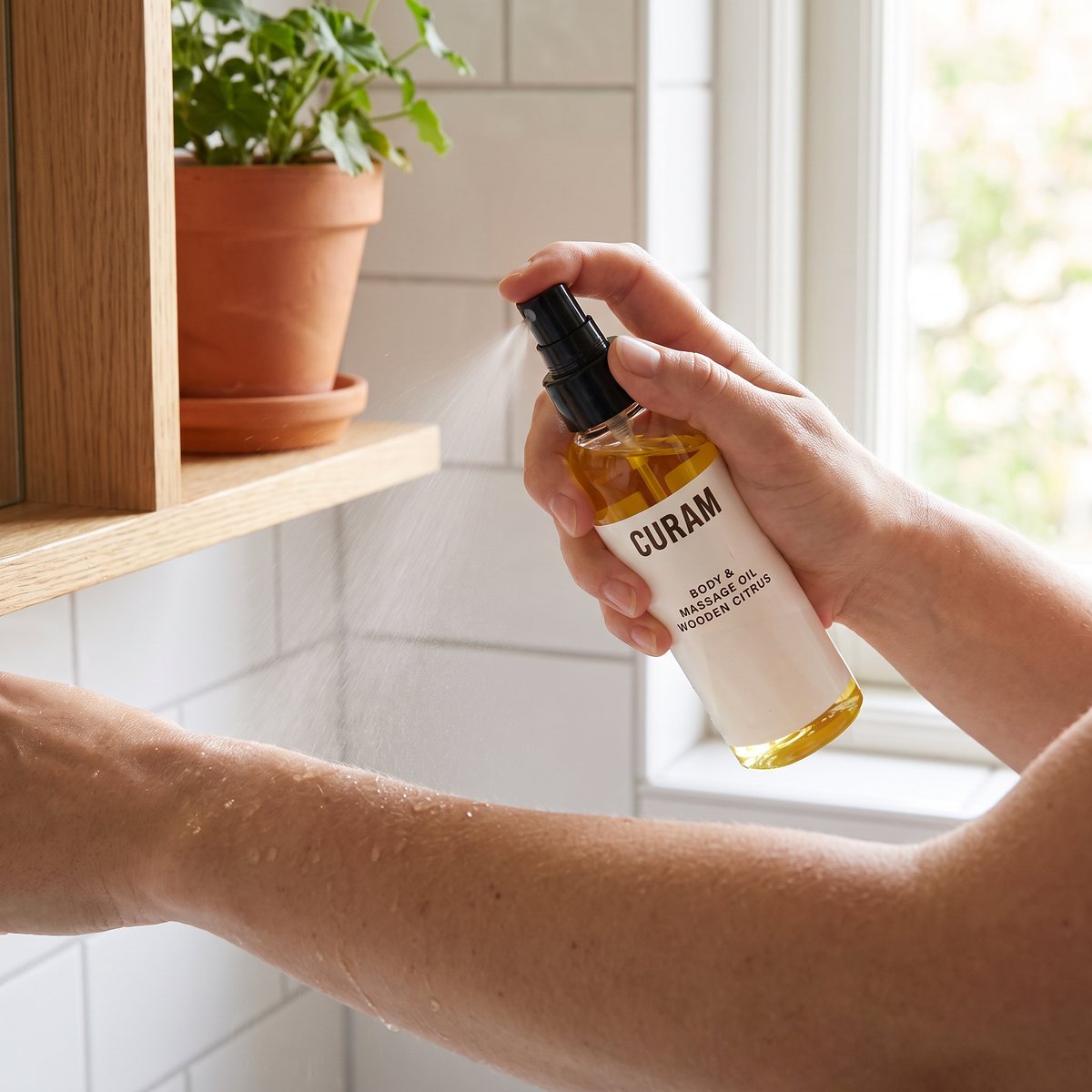 Hand som sprayar Curam massageolja på underarmen i ett ljust svenskt badrum med vitt kakel och en kruka