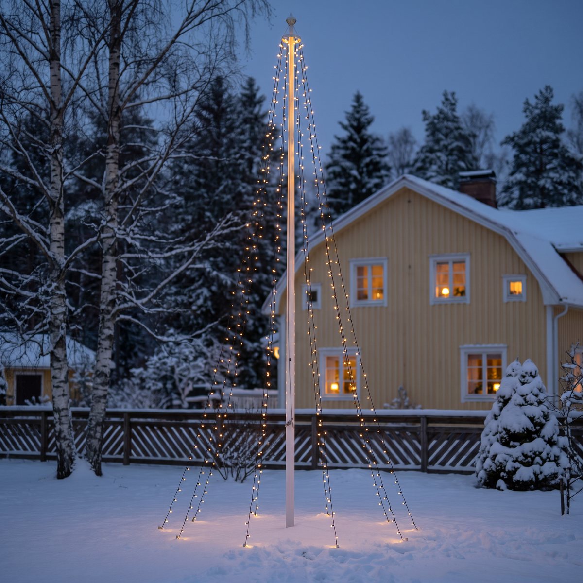 Flaggstång med varmgul ljuskon framför ett gult trähus och snöiga björkar en vinterkväll