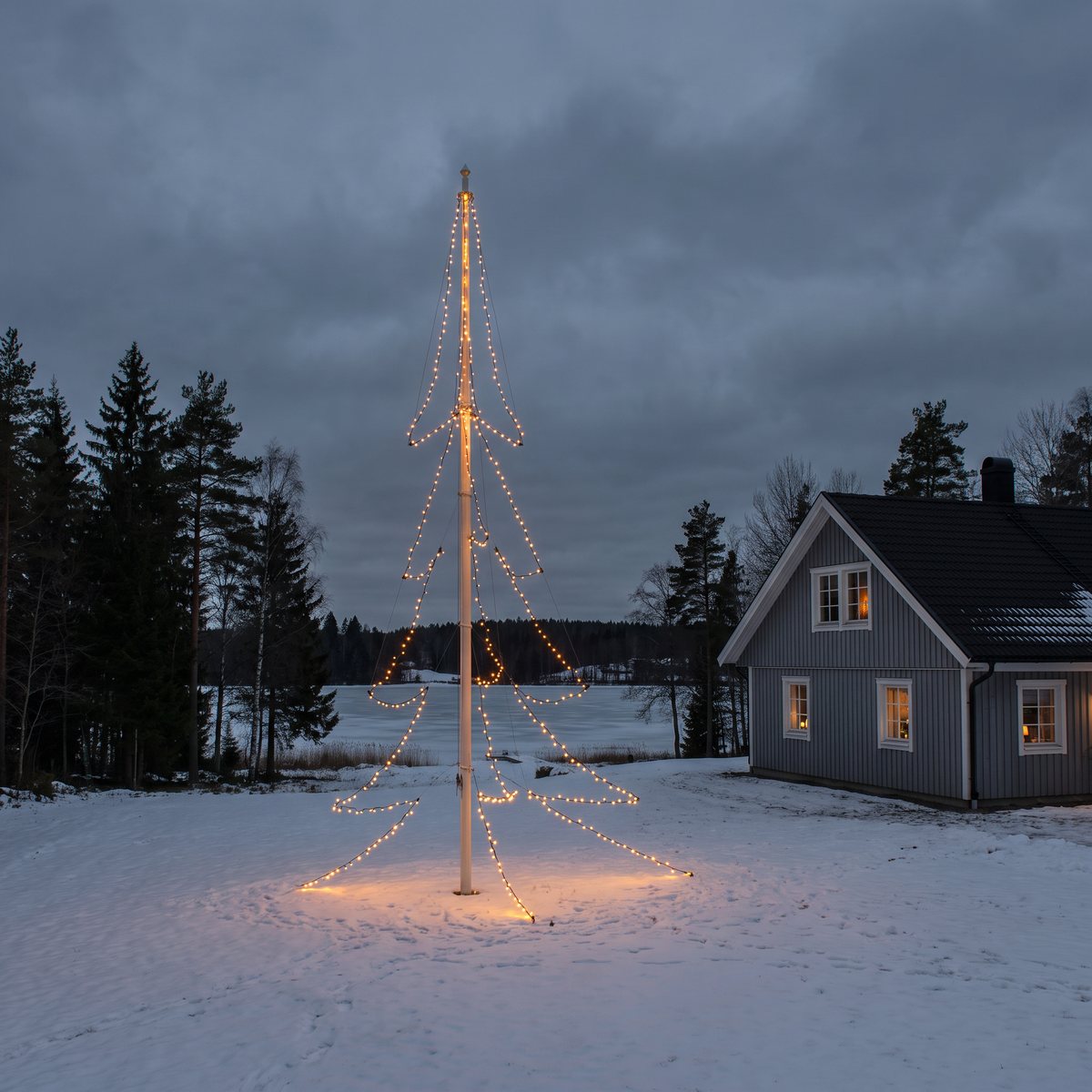 Hög flaggstång med amberfärgade ljusslingor formade som en gran på en snötäckt tomt vid en frusen sjö