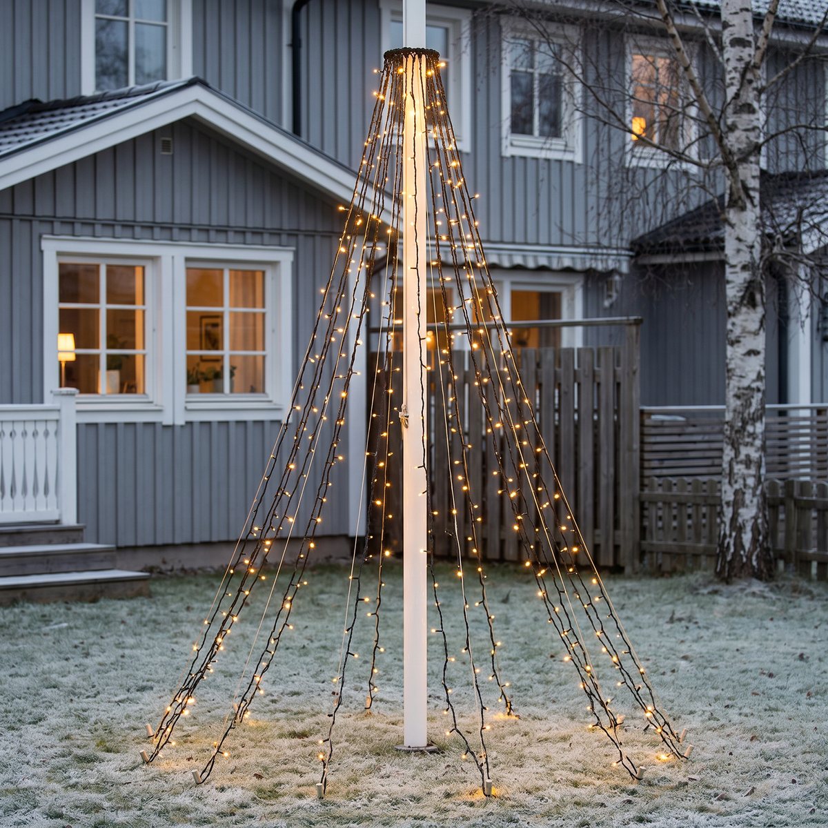 Lägre flaggstång med gyllene ljuskon på en frostig gräsmatta framför ett grått trähus