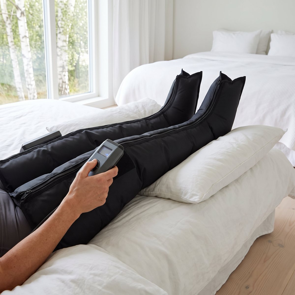 CurrentBody Wellness kompressionsstövlar på en person i ett ljust svenskt sovrum, kontrollenheten i handen