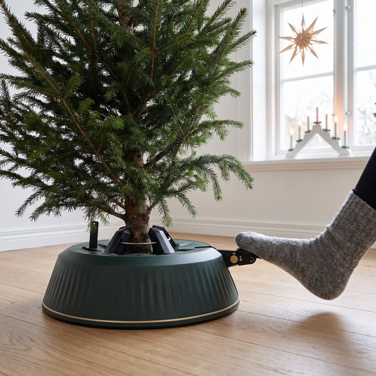 Mörkgrön Krinner X-240 julgransfot med en äkta gran, en fot i ullsocka trycker på fotpedalen på ett ljust trägolv i ett svenskt vardagsrum