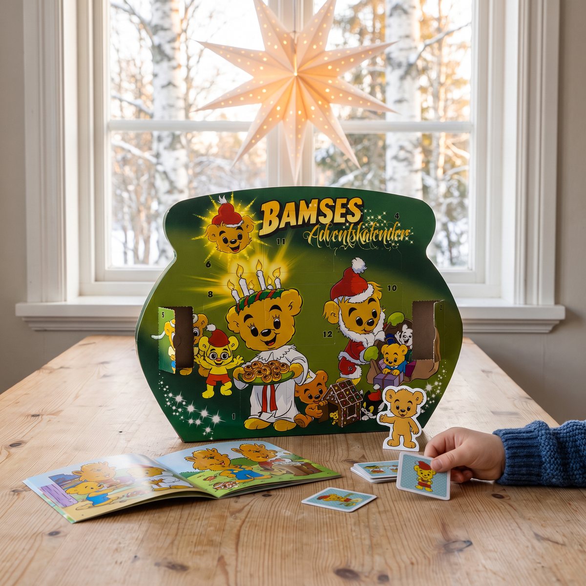 Bamses adventskalender i grön kartong på ett köksbord av furu framför ett fönster med adventsstjärna, med ett uppslaget sagohäfte, memokort och en pappfigur framför.