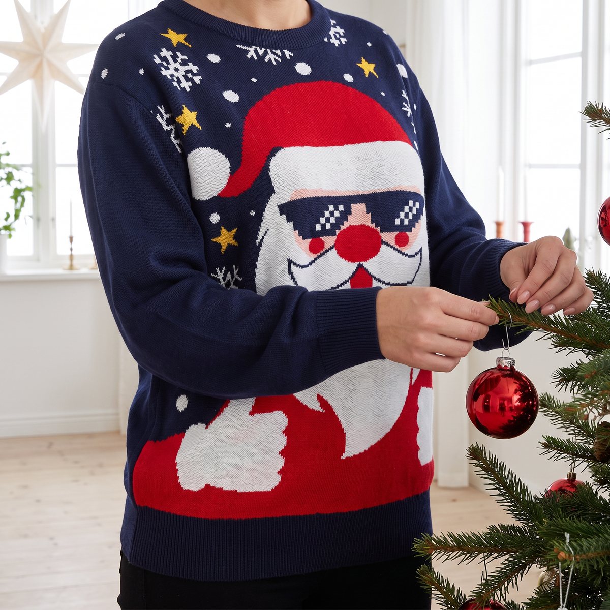 Kvinna i mörkblå jultröja med tomte i solglasögon hänger en röd julgranskula i granen i ett ljust svenskt vardagsrum