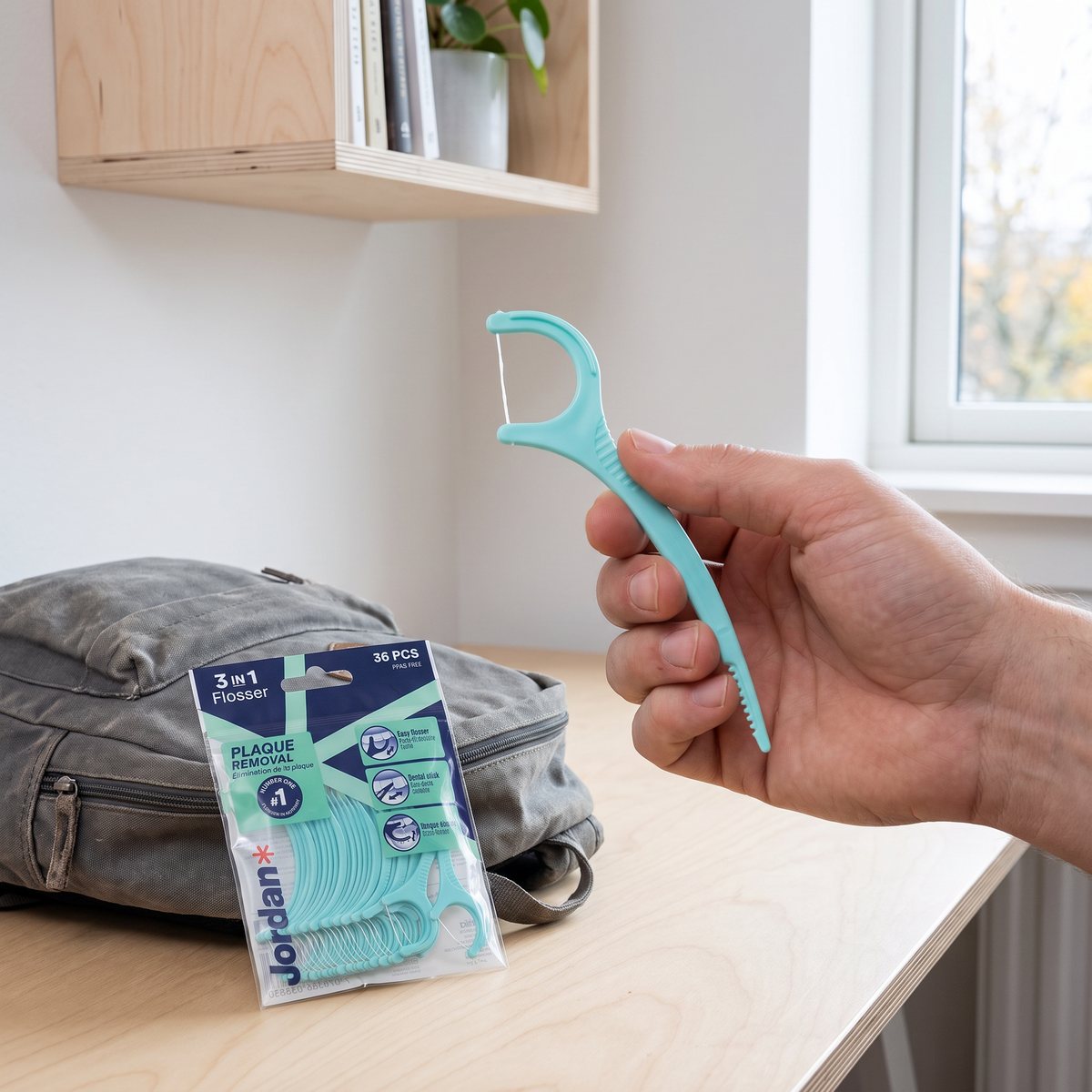 En hand håller en turkos Jordan 3-in-1 Flosser vid en transparent påse med fler byglar på ett träskrivbord i en svensk studentlägenhet.