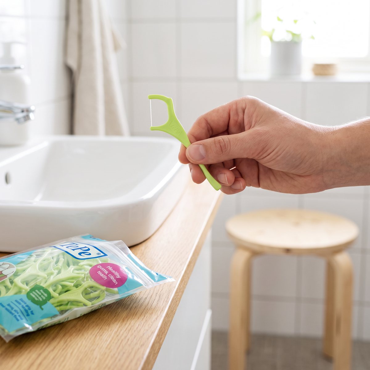 En hand håller en limegrön TePe Mini Flosser över handfatet i ett ljust svenskt badrum, påsen med tandtrådsbyglar på hyllan bredvid.