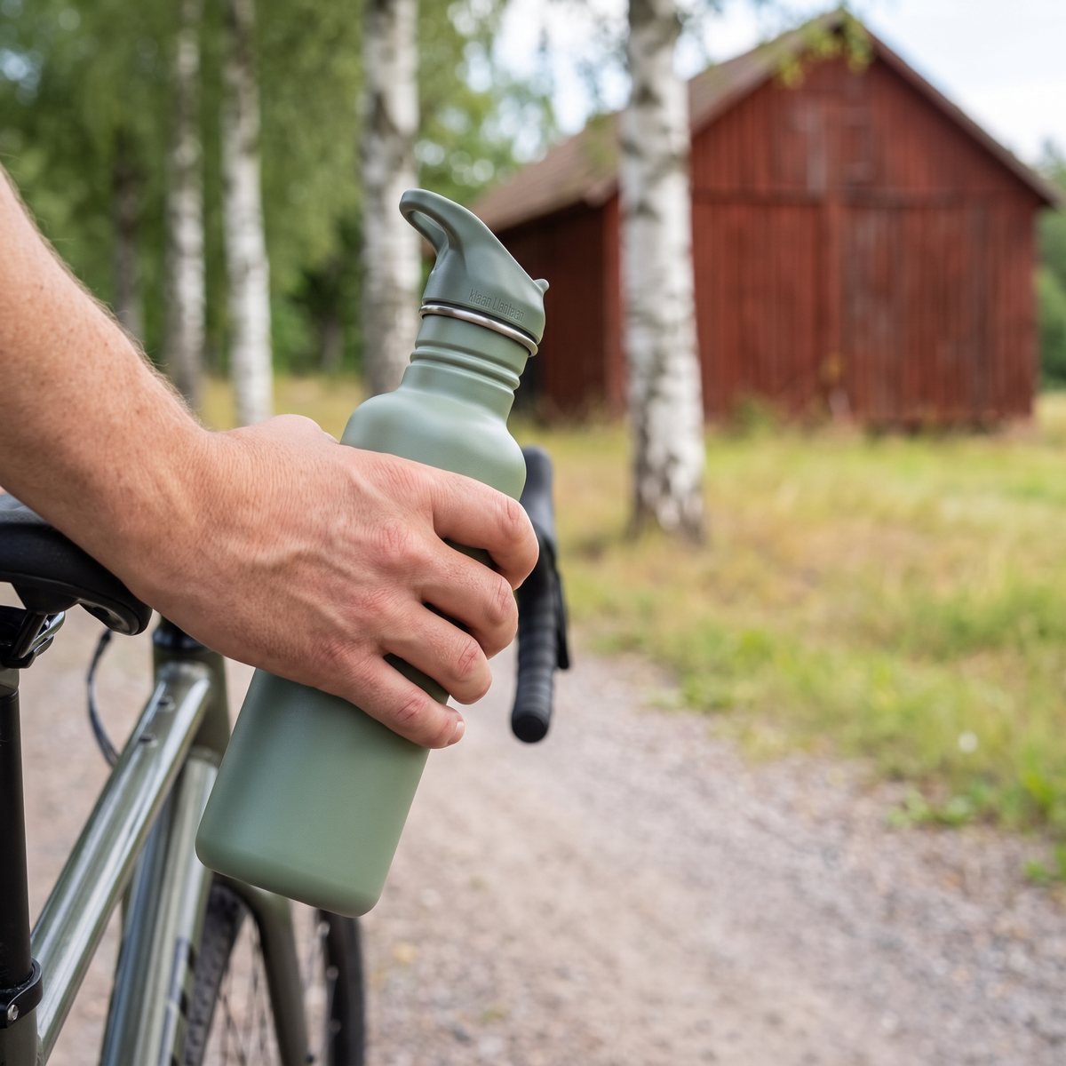 Klean Kanteen Classic i salviagrönt hållet i handen vid en grusväg på svensk landsbygd