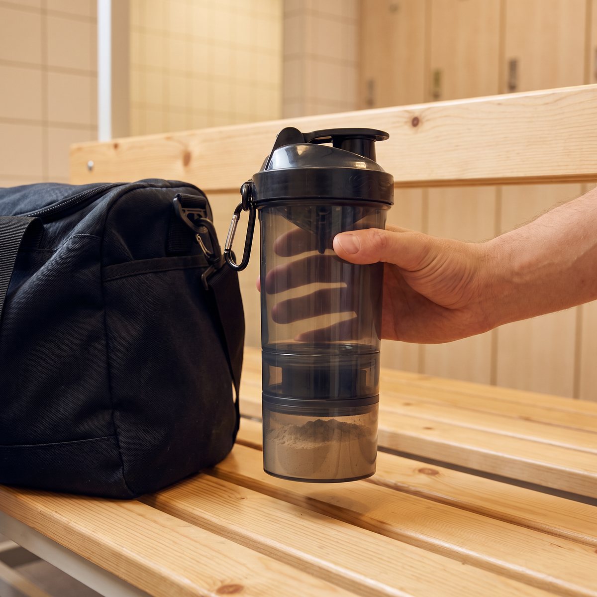 Smartshake Original2GO med proteinpulver i bottenfacket och karbinhake vid en träningsväska på en bänk i ett omklädningsrum