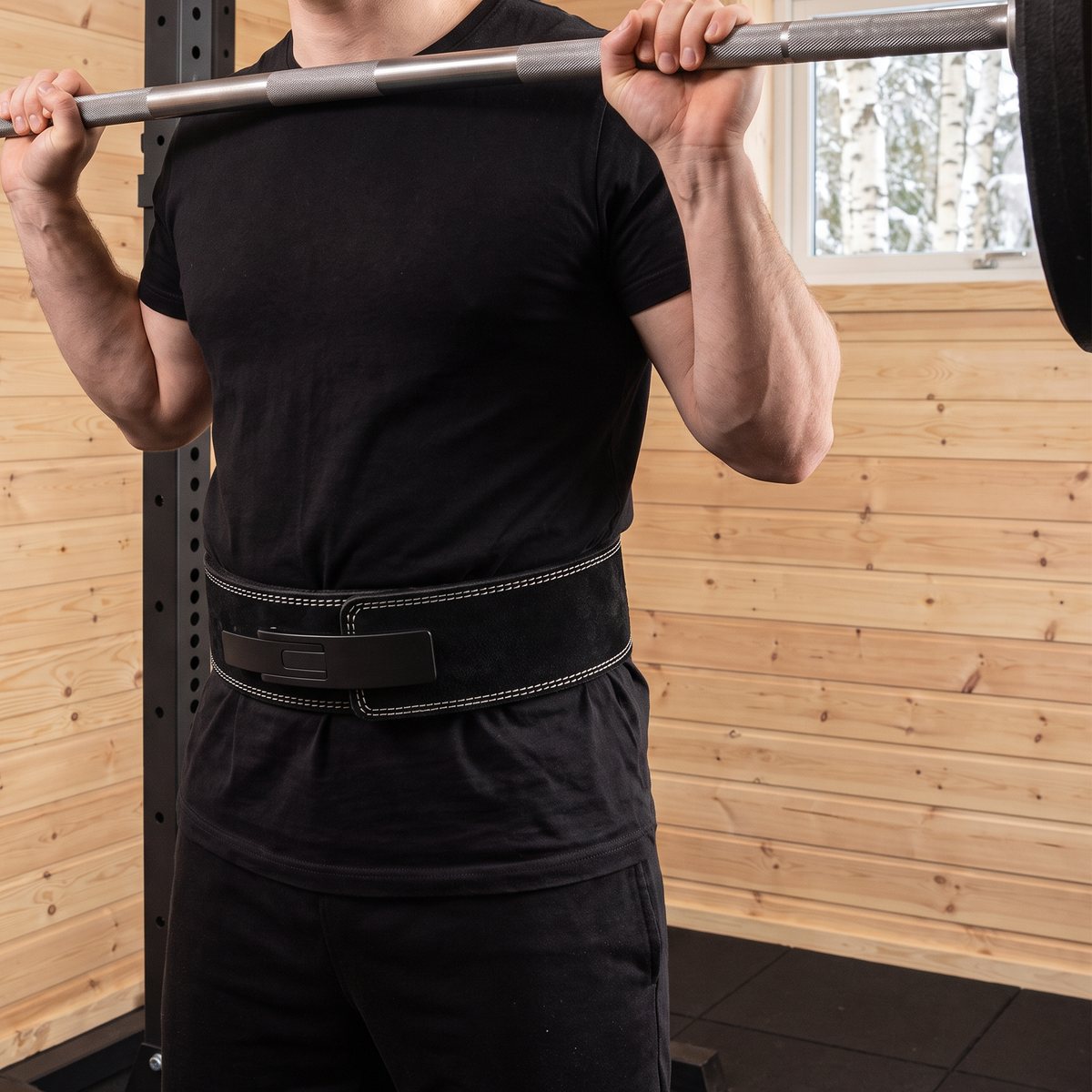 C.P. Sports Powerlifting Lever Belt i svart mocka med hävstångsspänne runt midjan på en lyftare vid skivstången i ett svenskt källargym med panelväggar och snöiga björkar utanför