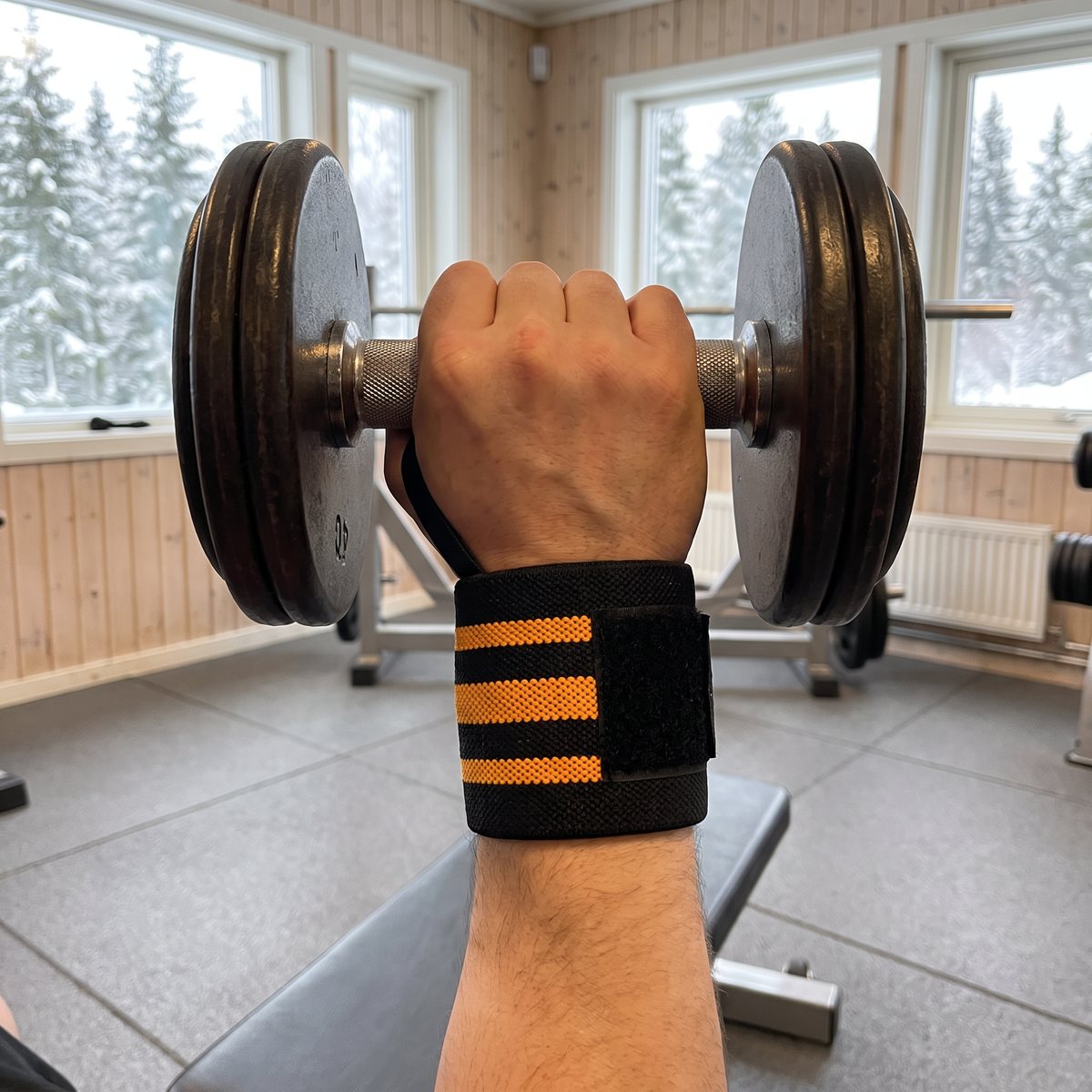 Svarta handledsstöd med orangea ränder på en handled i ett svenskt gym under hantelpress