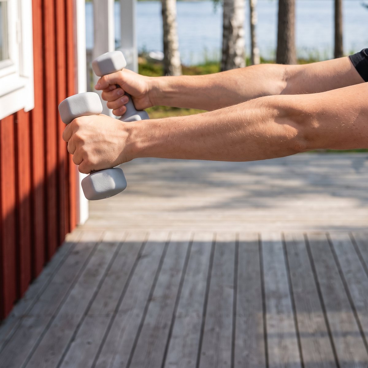 Två ljusgrå Abilica neoprenhantlar i framåtlyft på altanen till en röd svensk sommarstuga vid en sjö