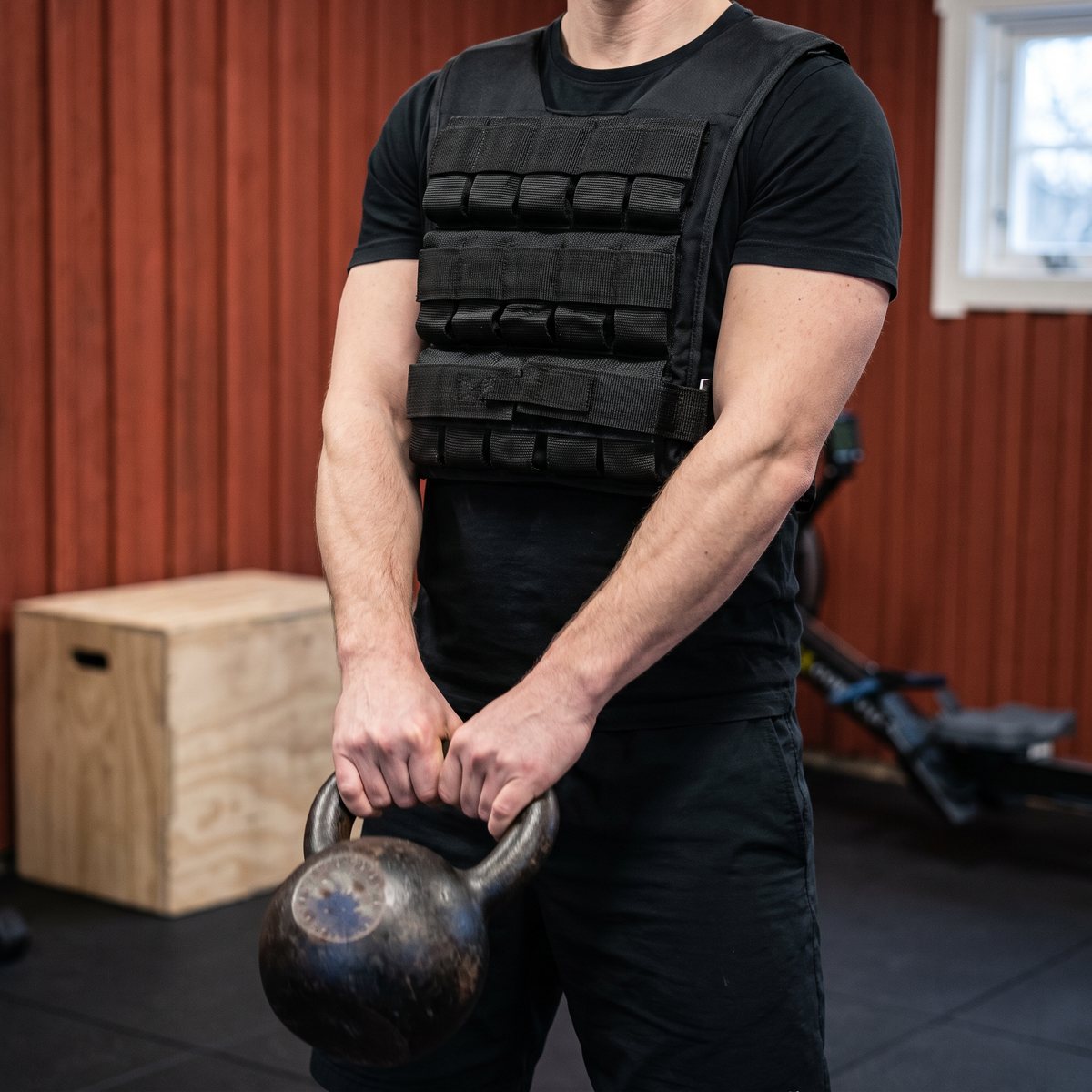 Svart viktväst från Toorx buren under kettlebellträning i ett garagegym med röd träpanel, gummigolv, plyobox och roddmaskin.