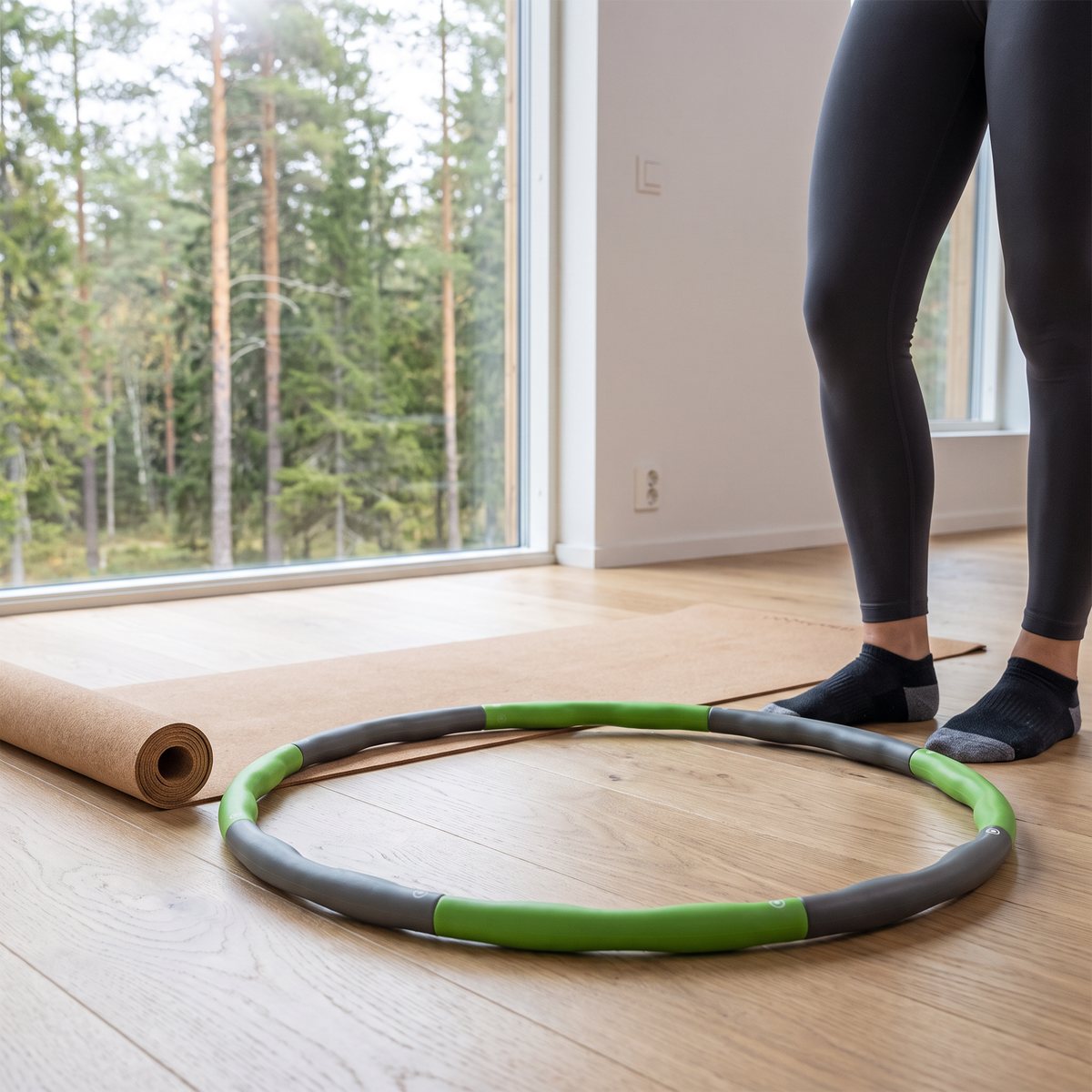 Grön och grå delbar hulahoppring liggande på trägolv bredvid en yogamatta i en svensk lägenhet med tallskog utanför fönstret