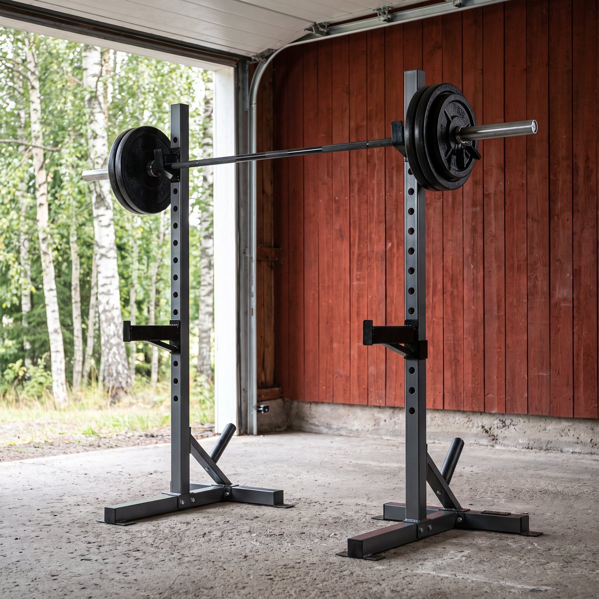 Fristående squatställning med lastad skivstång i ett svenskt garage med röd träfasad och björkar