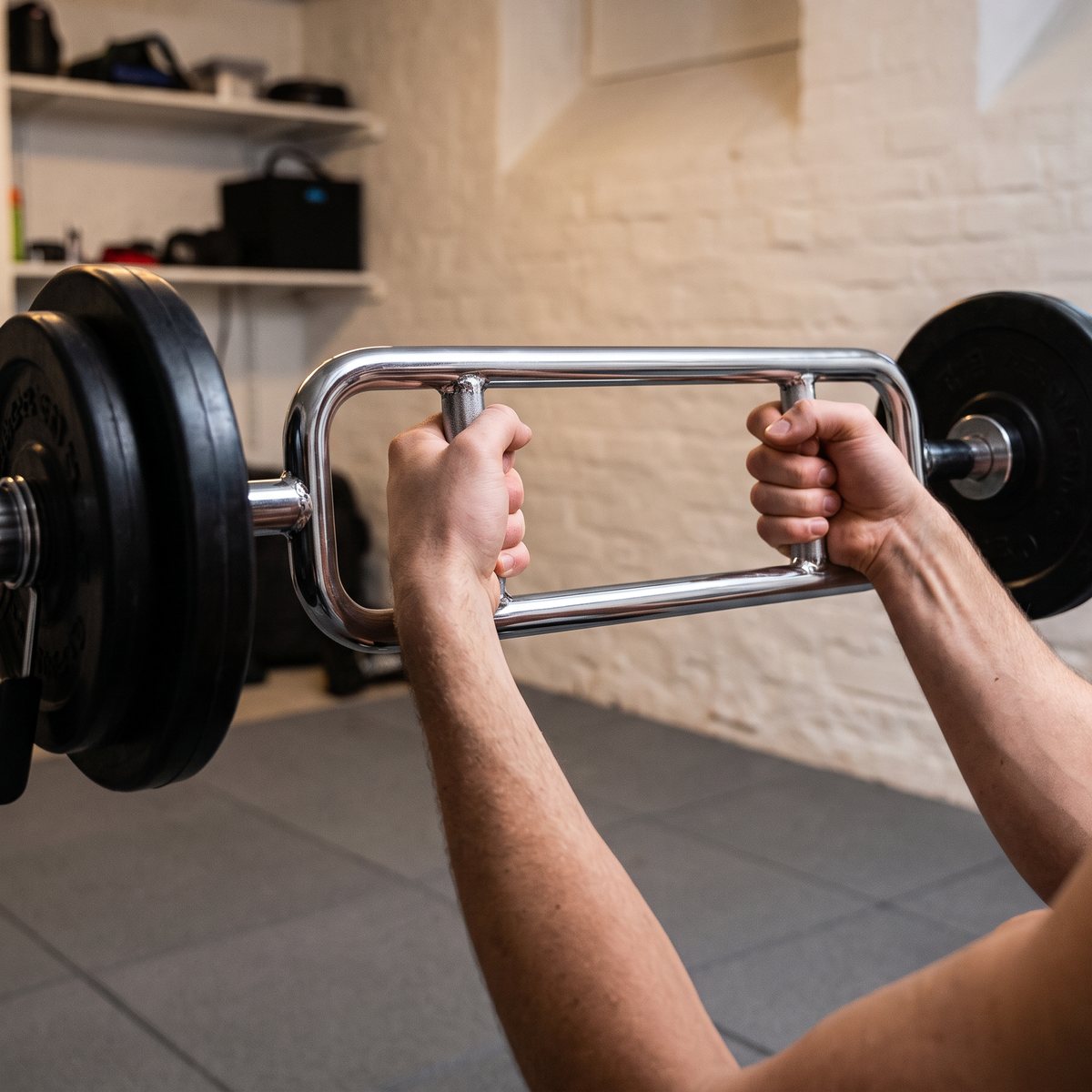 Kromad tricepsstång med lastade skivor hålls i bröstnivå i en svensk källargym med vitkalkade tegelväggar