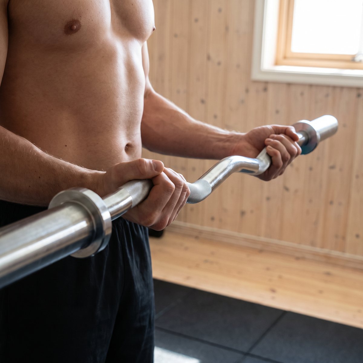 Master Fitness curlstång i krom hålls med underhandsgrepp vid en bicepscurl i ett svenskt hemmagym i källaren med furupanel