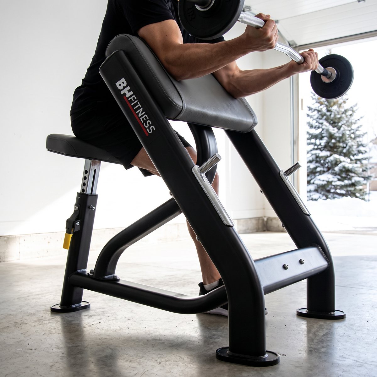 BH Fitness L830BB i ett ljust svenskt garagegym med snötäckt gran utanför, där någon curlar en skivstång mot det vinklade armstödet