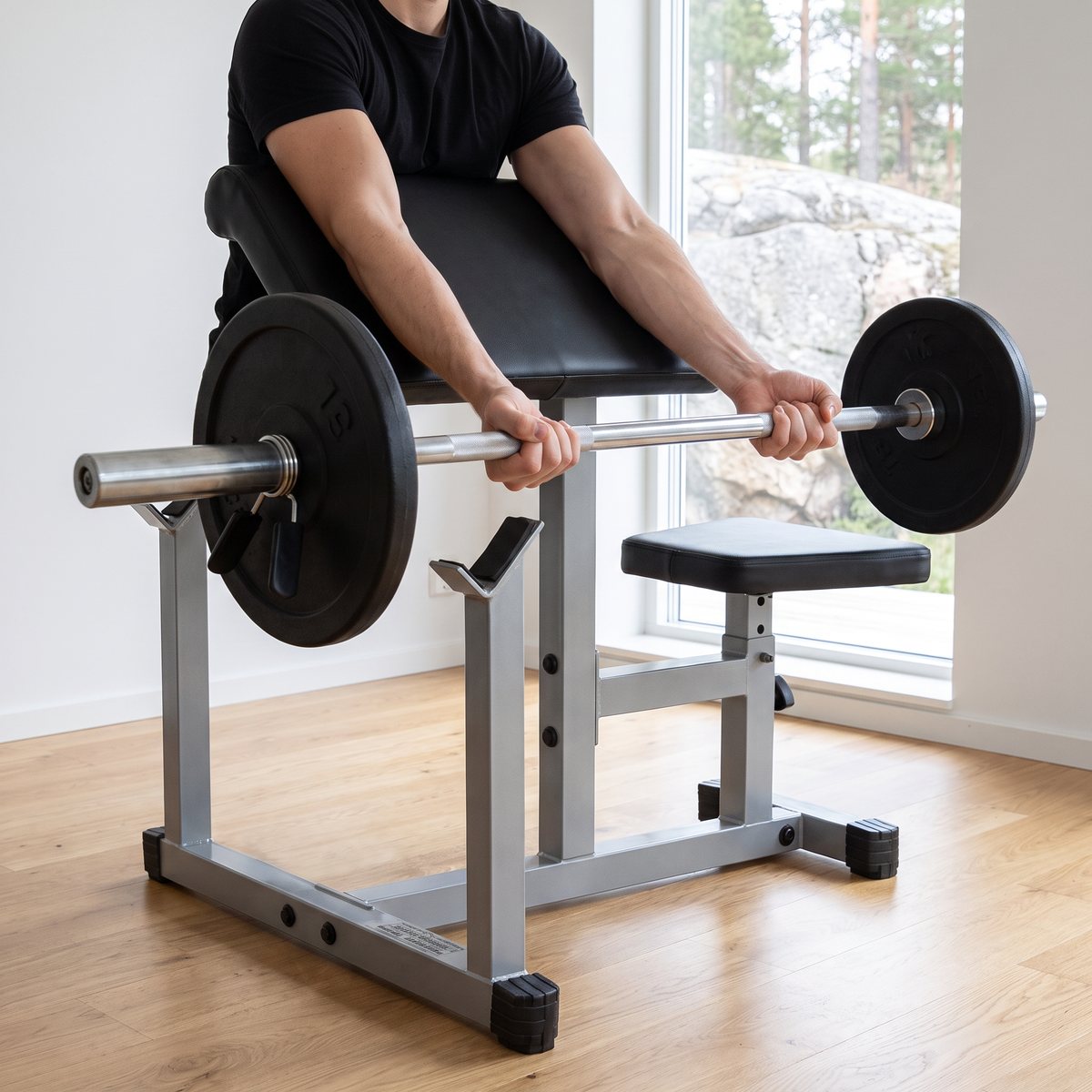 Body Solid PPB32 i ett vitt svenskt gymrum med trägolv och granitberg utanför fönstret, där någon curlar en skivstång mot det stoppade armstödet