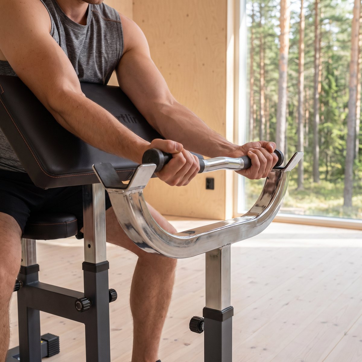 inSPORTline Scott Curl Bench LKC301 i ett svenskt loftgym med ljusa björkplywoodväggar och tallskog utanför fönstret, där någon curlar en stång mot det stoppade armstödet