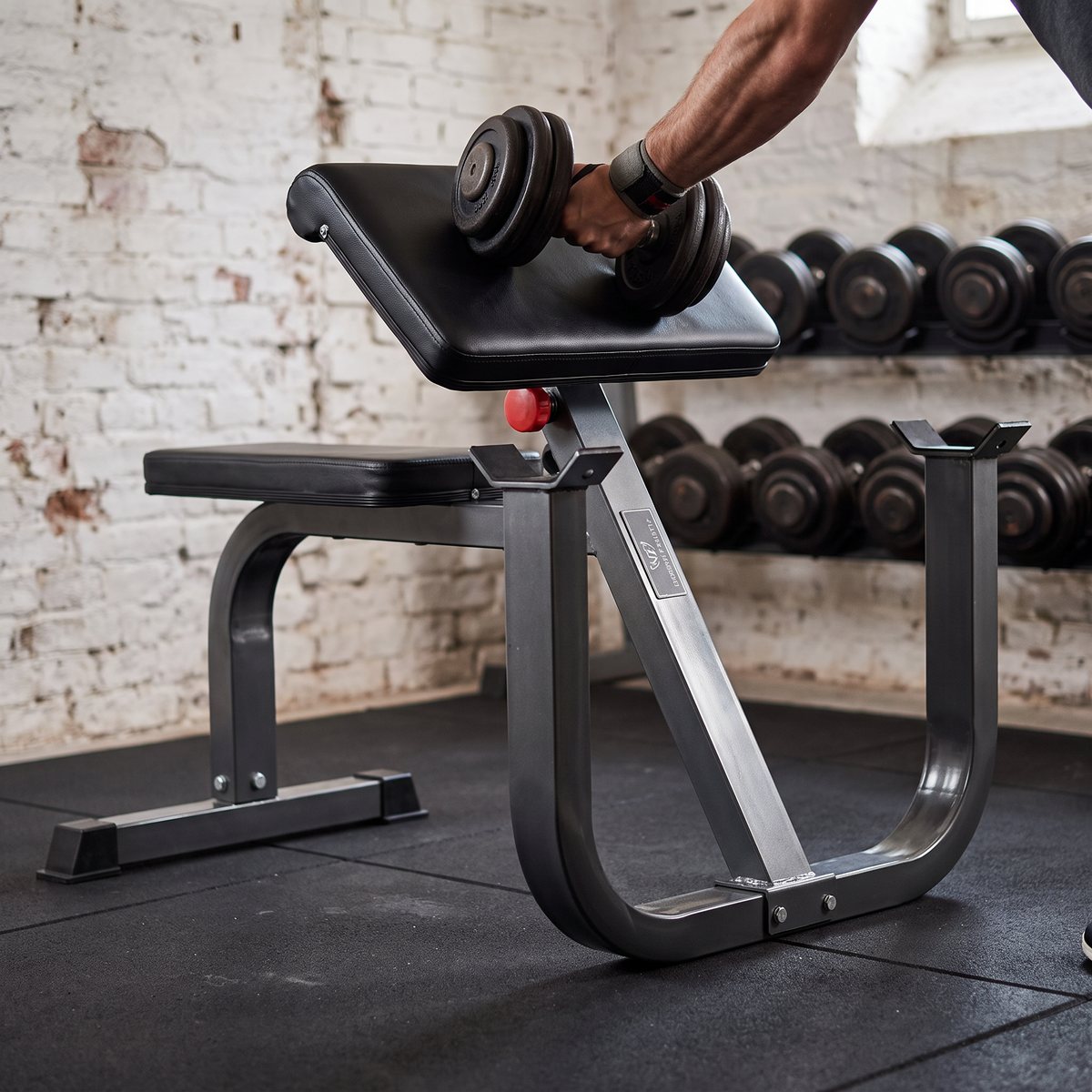 Nordic Fighter Preacher Curl i ett källargym med vitkalkade tegelväggar och hantlar i bakgrunden, där en hand håller en hantel mot det stoppade armstödet