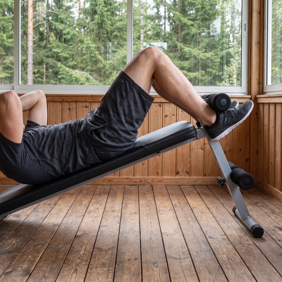 Everfit WBK-100, en grå och svart magbänk, används för situps på en svensk glasveranda med plankgolv och granskog utanför
