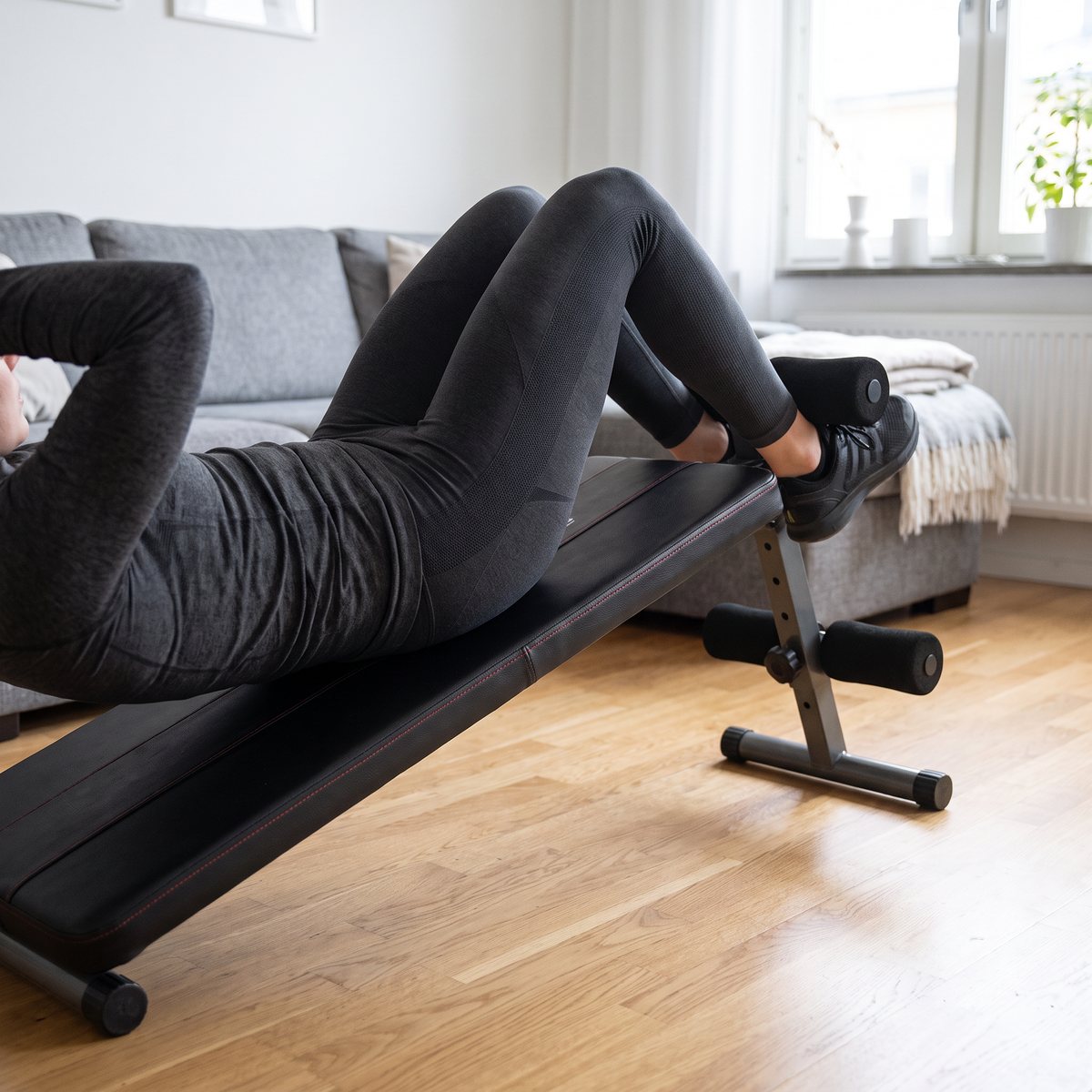 inSPORTline Situpsbänk, en svart situpsbänk, används för situps i ett svenskt vardagsrum med ekparkett