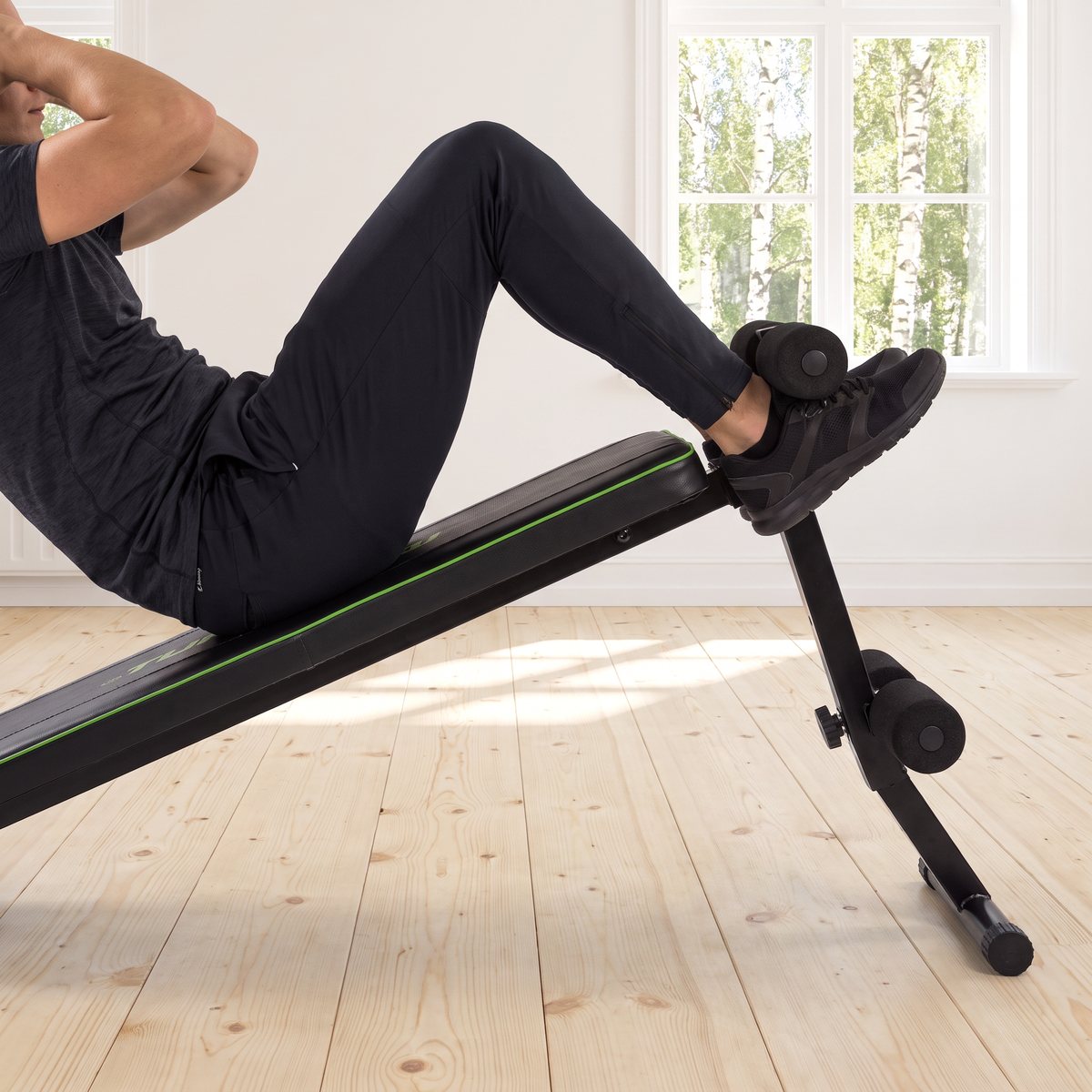Tunturi AB20 Abdominal Bench, en svart magbänk med grön kant, används för crunches i ett ljust svenskt vardagsrum med furugolv